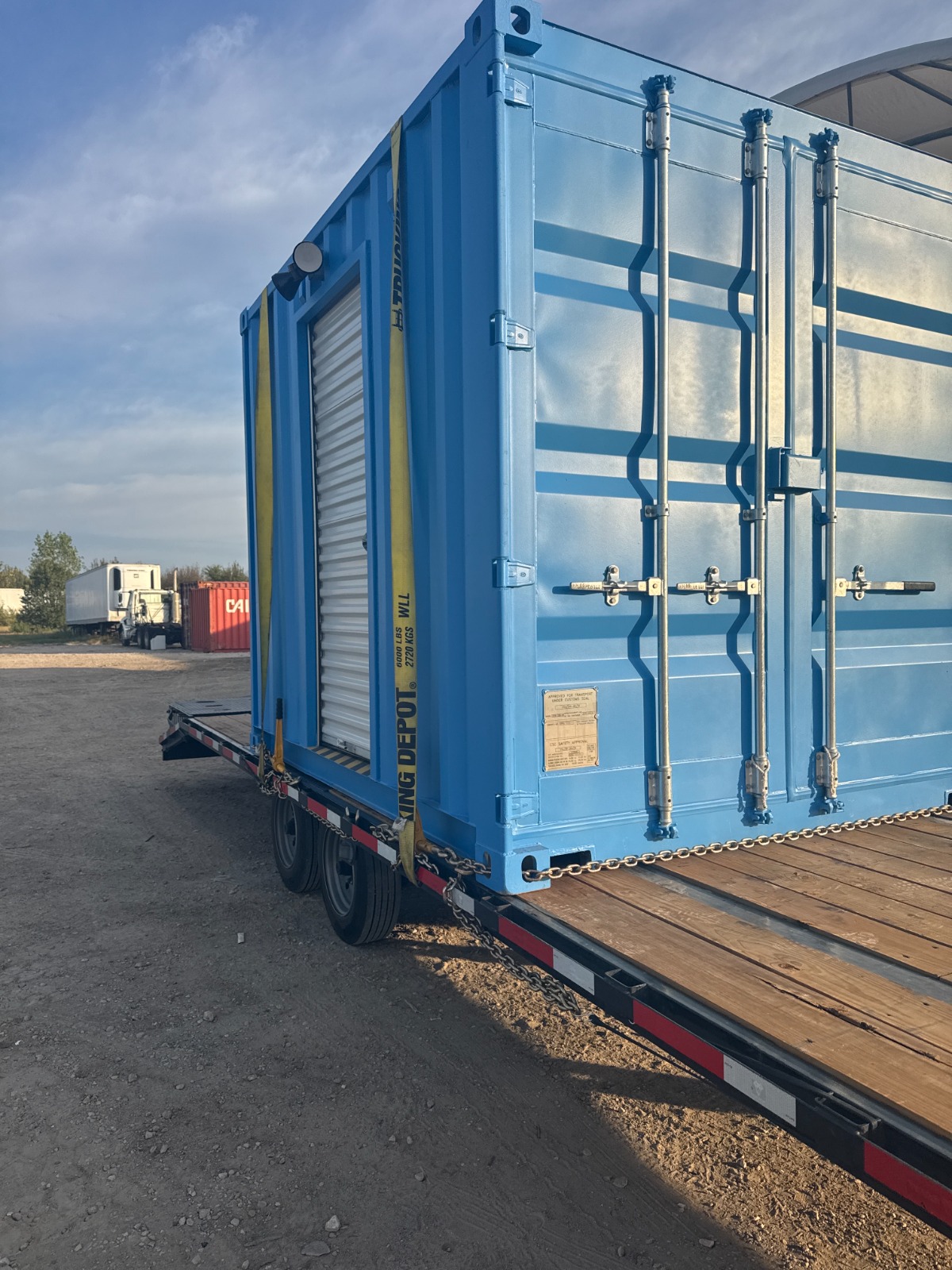 A blue 10-foot shipping container turnstile checkpoint secured with chains and straps on a wooden flatbed trailer