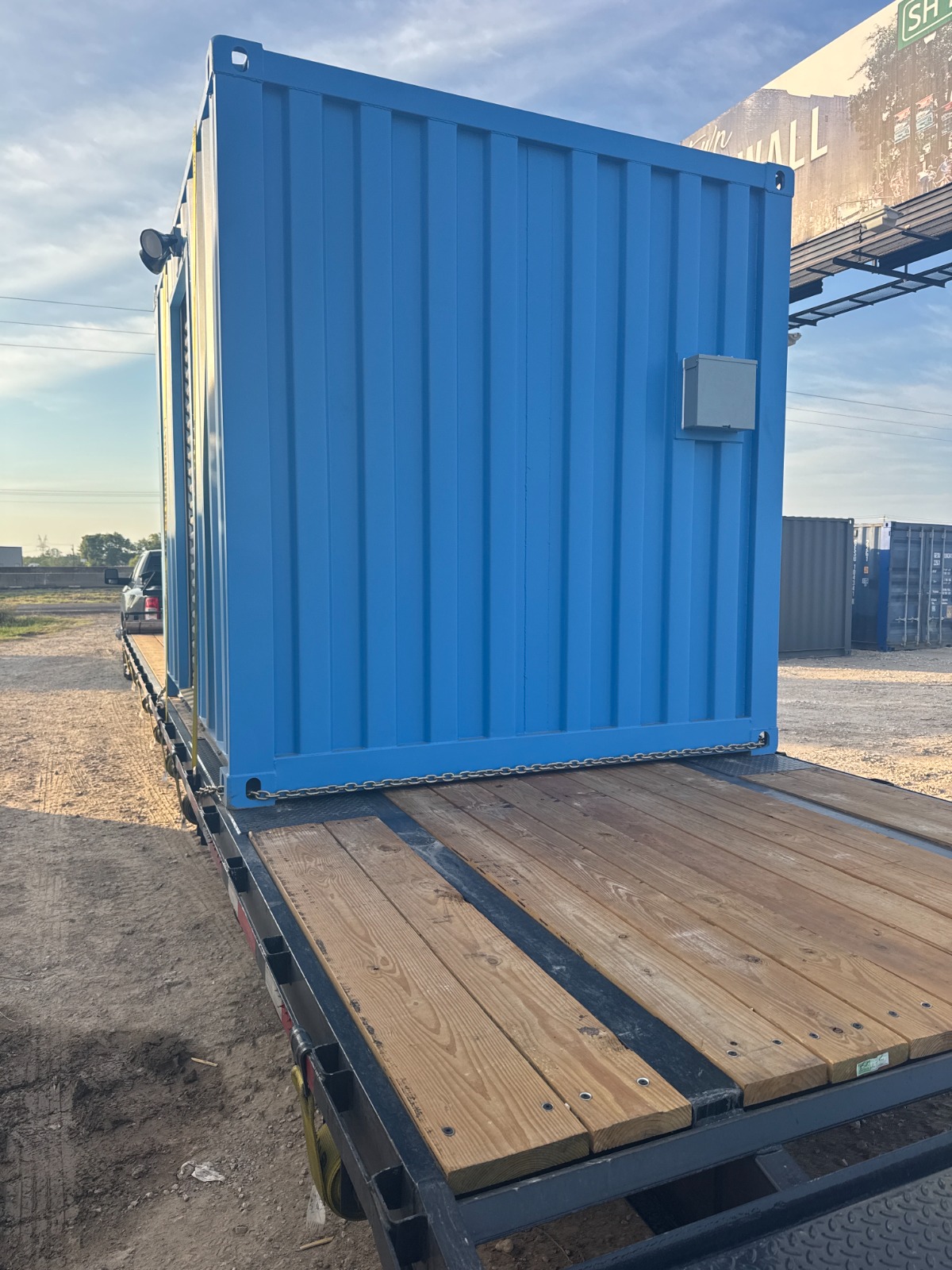 A blue 10-foot shipping container turnstile checkpoint with a gray electrical panel secured with chains on a wooden-deck flatbed trailer