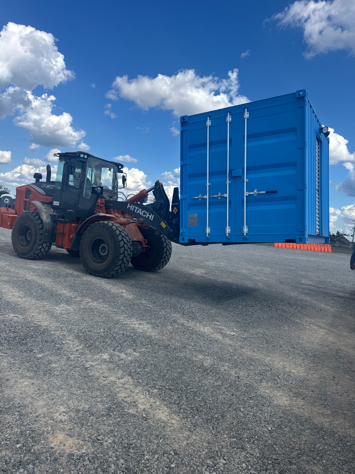 A wheel loader lifts a blue 10-foot shipping container turnstile checkpoint across a gravel yard