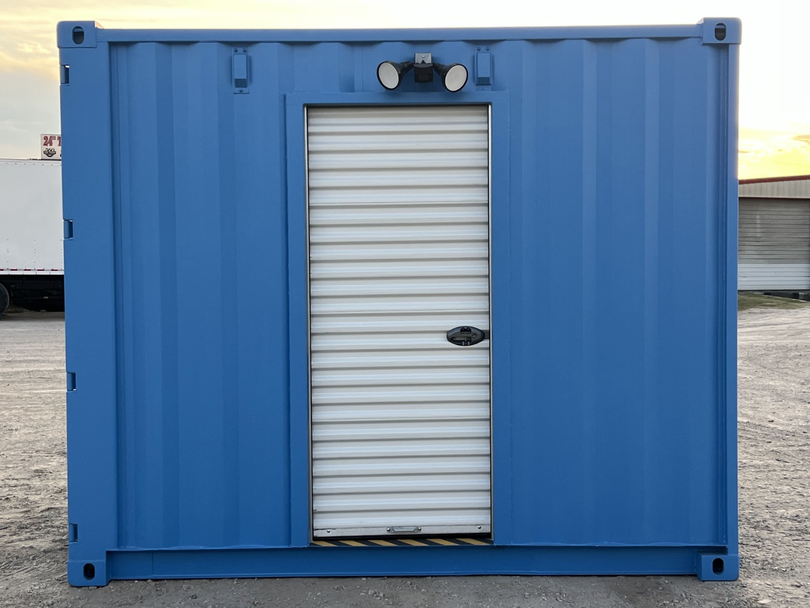 Straight-on exterior view of a blue 10-foot shipping container turnstile checkpoint with a closed white roll-up security door and overhead floodlight