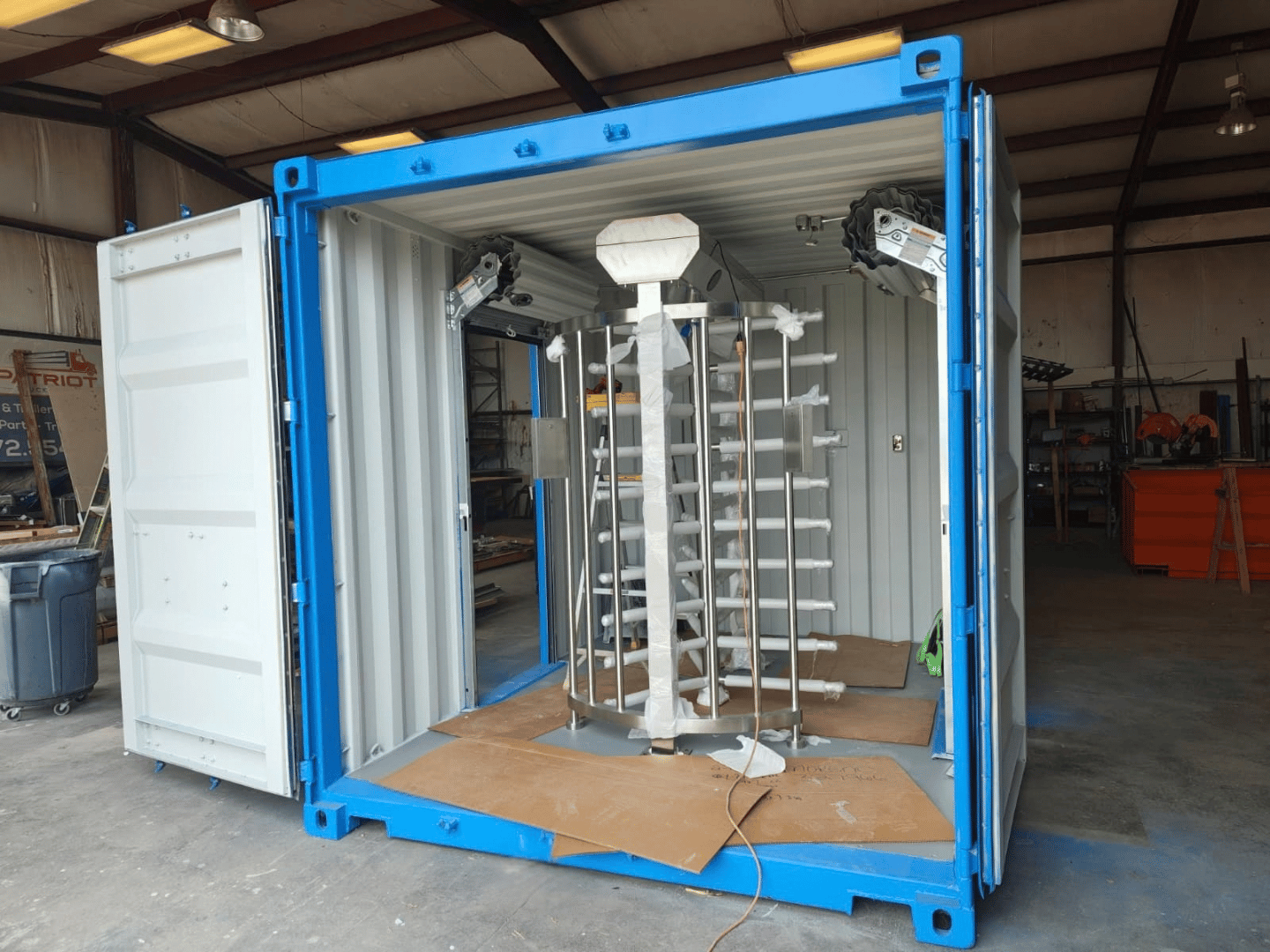 Wide-angle interior view of a full-height stainless steel turnstile installed inside a 10-foot shipping container checkpoint with roll-up side doors